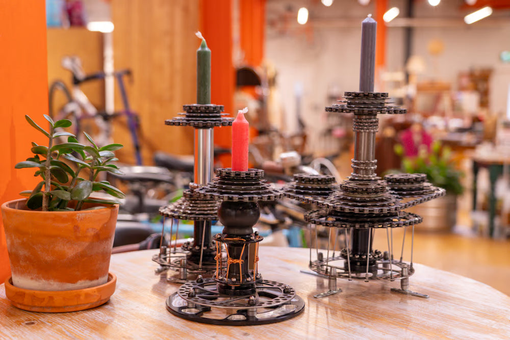 Two decorative metal candle holders with a gear-like design, styled on a wooden table in a warm, blurred shop environment. One holder is black with a green candle, and the other is silver with a red candle, next to a potted plant.