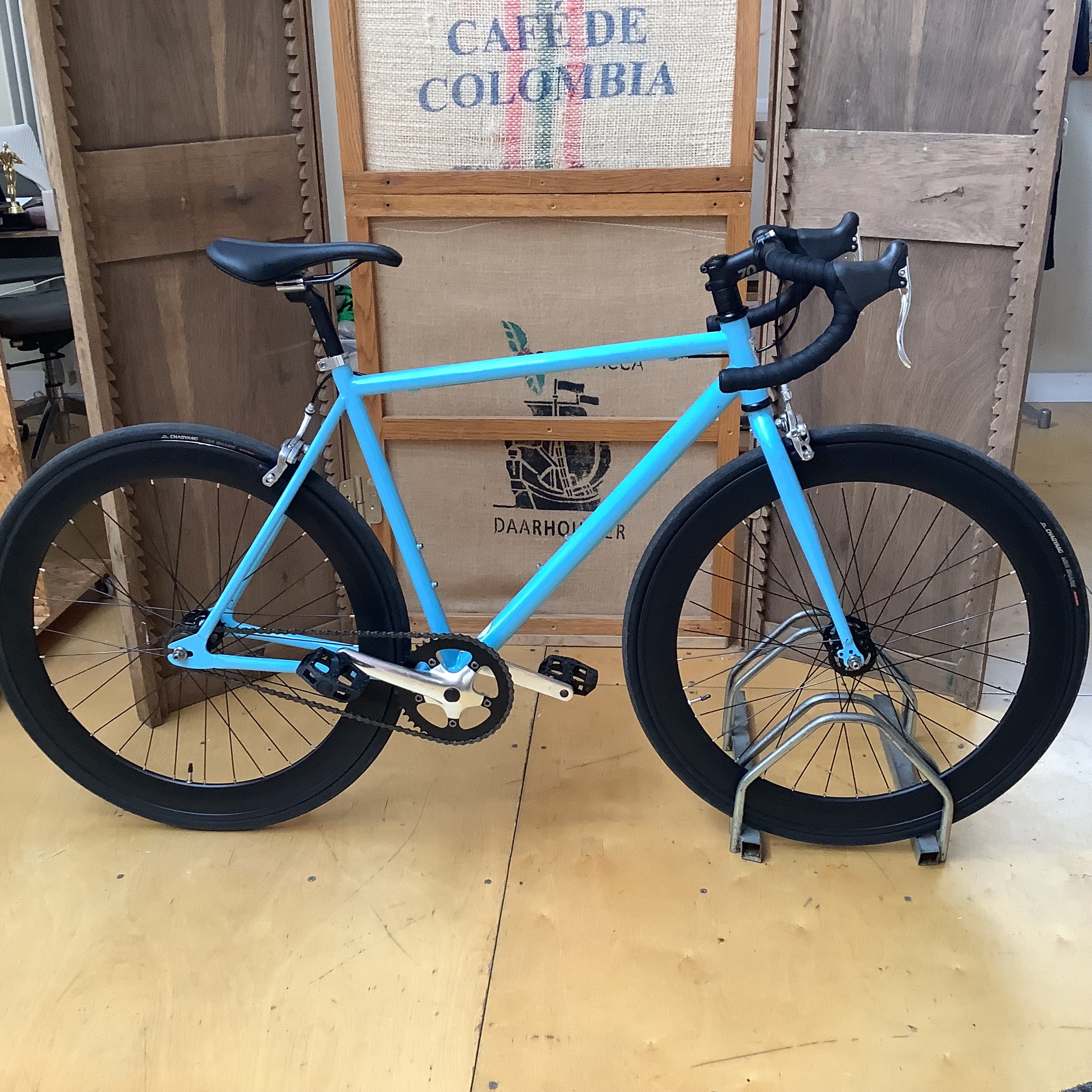 A bright turquoise road bicycle with black components, displayed on a metal stand in front of wooden coffee roaster crates.