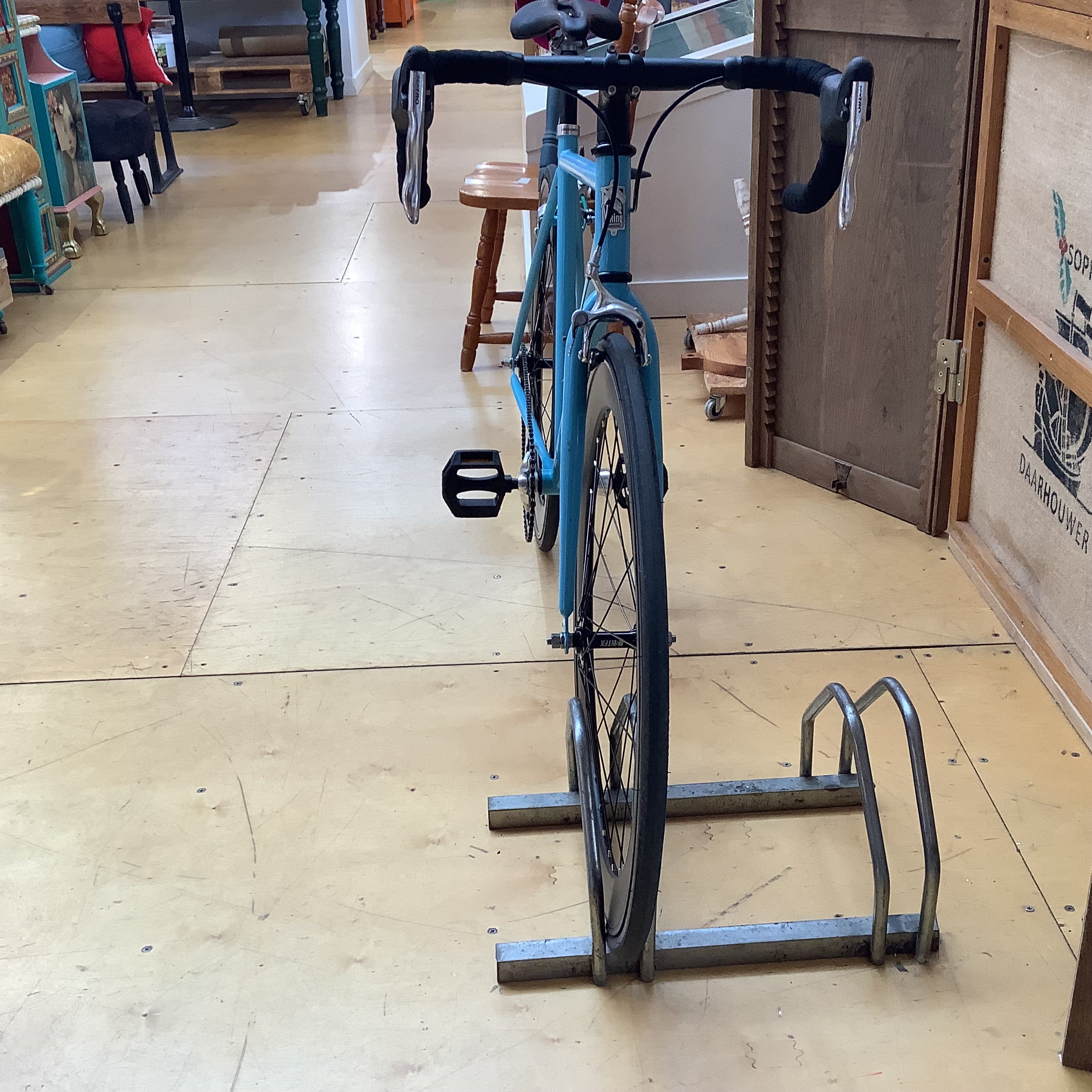 A turquoise road bicycle with black handlebars and tires is parked on a metal bike rack in a shop with a light wood floor.