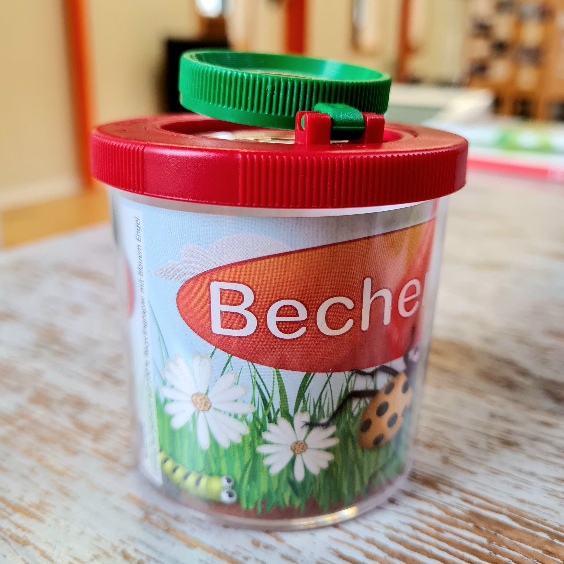 A large magnifying cup with a red and green lid, featuring a label with a bee and some daisies, and the brand 'Beco' on the front.