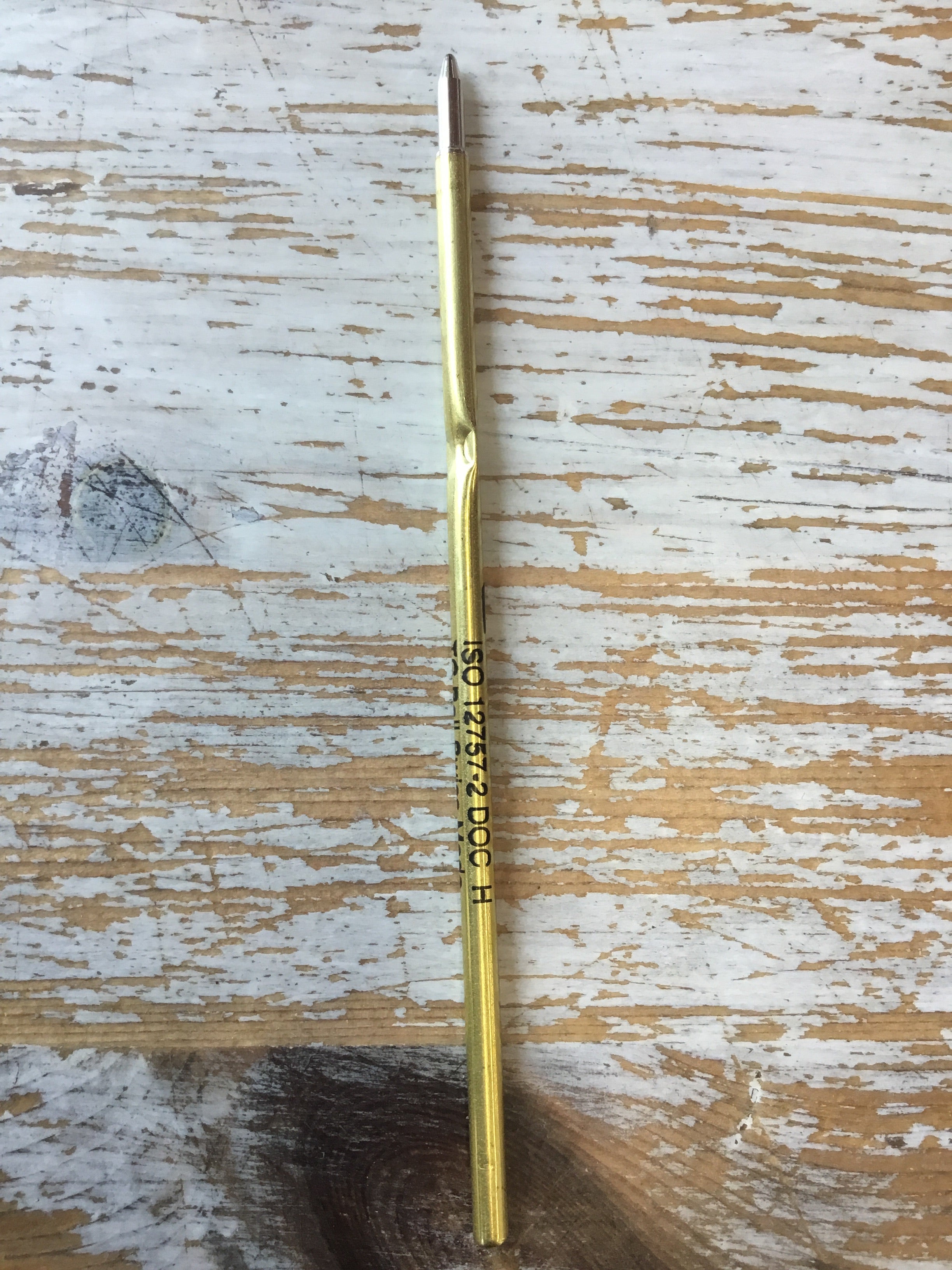 A gold-colored retractable wide nib biro refill lying on a wooden surface with visible wood grain.