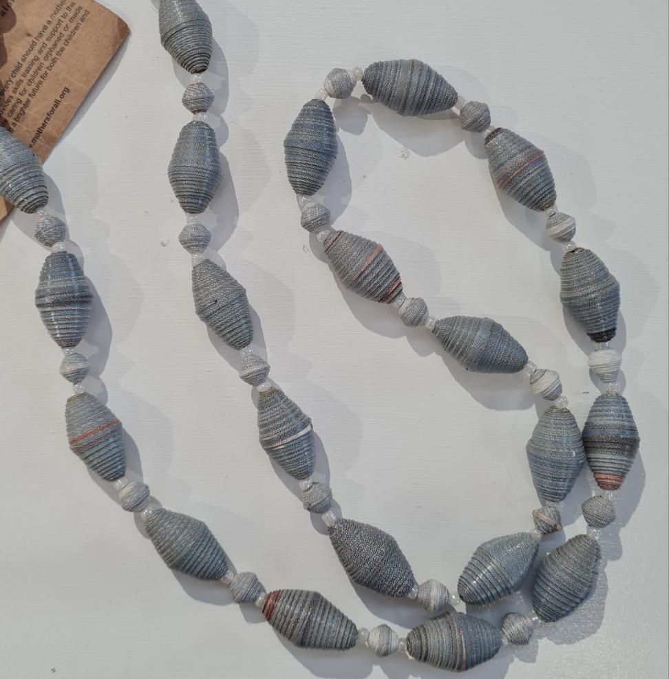 A long necklace with white, grey, and blue recycled magazine beads laid out on a white surface.